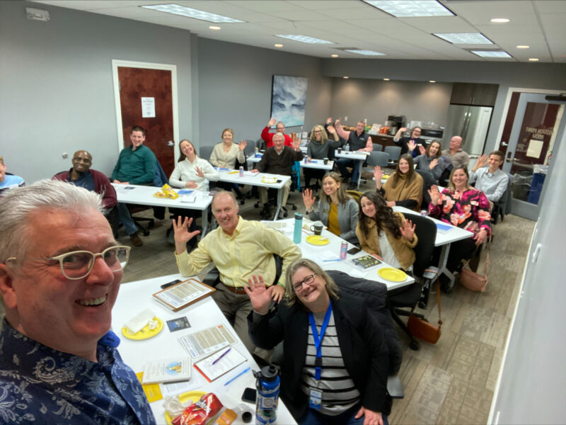 A group of people sit at angled tables in a conference room with one white man in the foreground. They are smiling and waving at the camera, posing for a photo during a workshop.