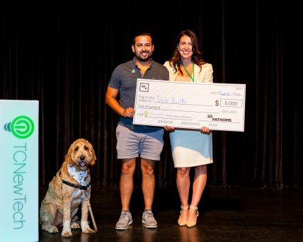 Two people stand on stage holding a giant check. A dog wearing a bandana sits next to them.