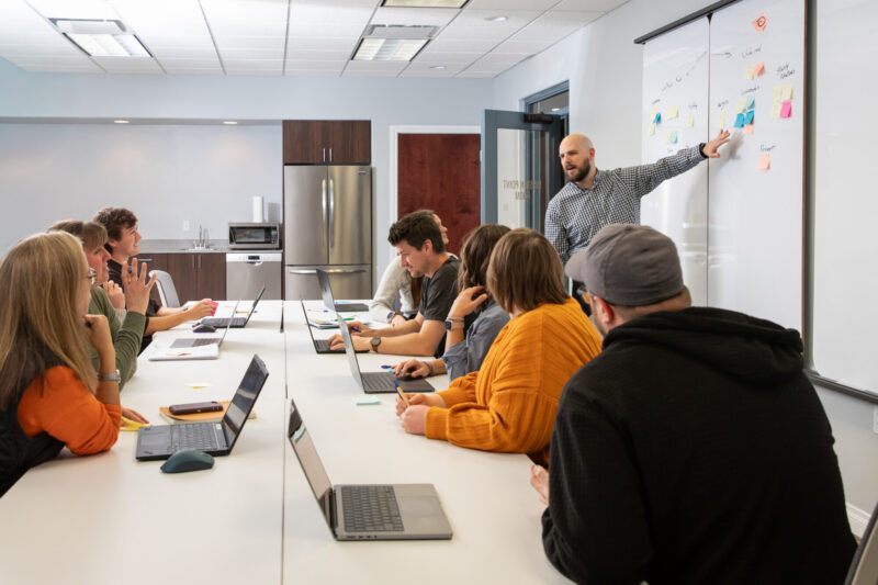 A bald white man with a beard gestures to the white board in a large conference room while others listen.