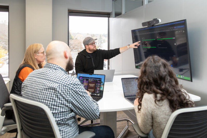 A white man wearing a gray baseball hat gestures towards a screen while others with laptops listen.