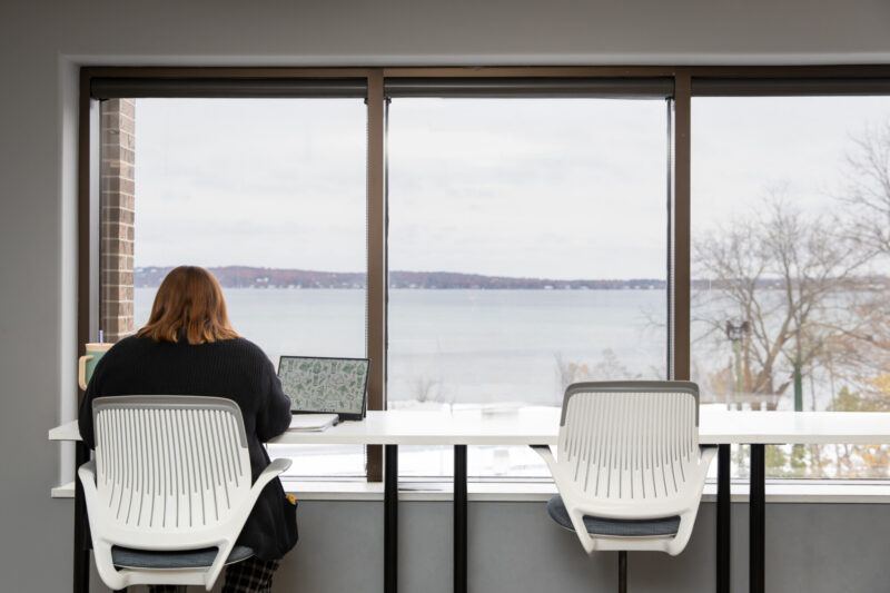 A person with long hair wearing a black sweater faces their laptop. They are sitting in front of the large windows at the 20Fathoms office overlooking the Bay.