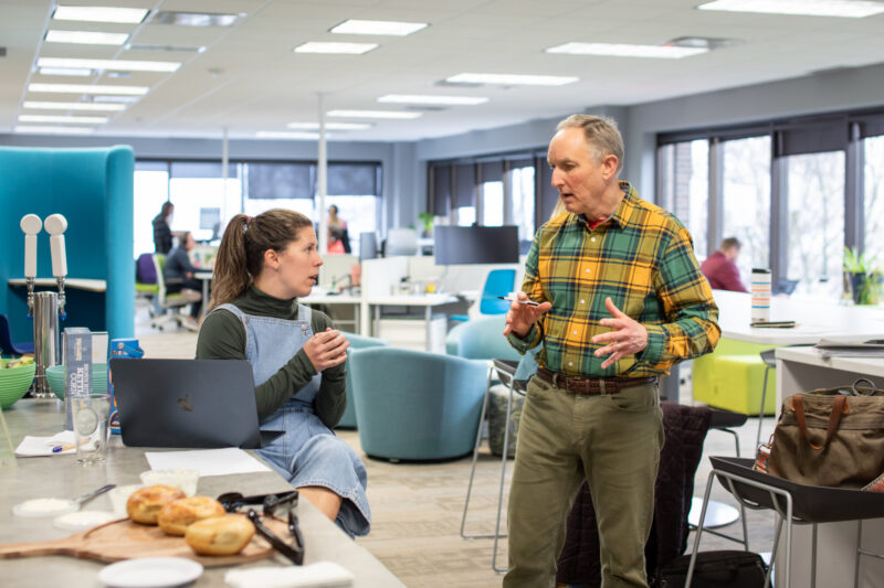A woman in overalls with a ponytail chats with a man in a plaid shirt at Breakfast Lab, a weekly educational event for entrepreneurs at 20Fathoms.